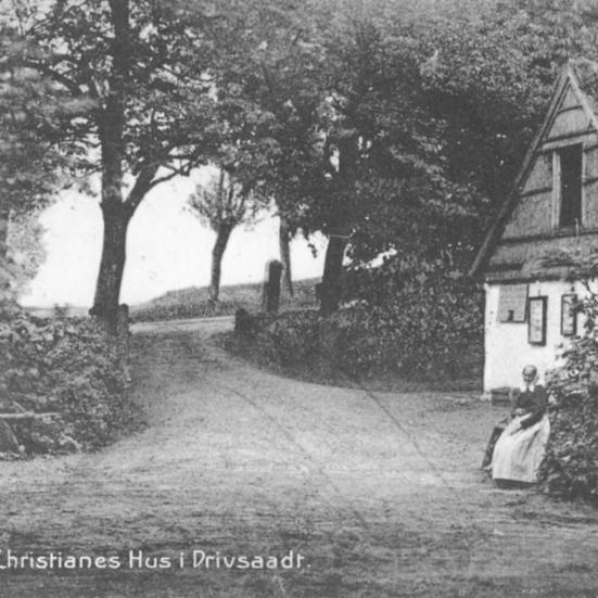 Sort-hvid foto: Facaden af Christianes Hus med Christiane, der sidder foran på en bænk.