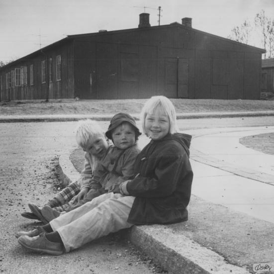 Sort-hvid foto af tre børn siddende på fortovet foran en sort træbygning.