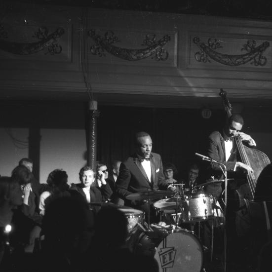 Sort-hvid foto af jazzband på scene med kontrabas, klaver og trommesæt. Dæmpet belysning og publikum uden om scenen.