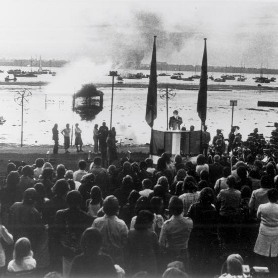 Sort-hvid foto af en menneskemængde foran en talerstol foran fjorden. Der brænder et bål på vandet.