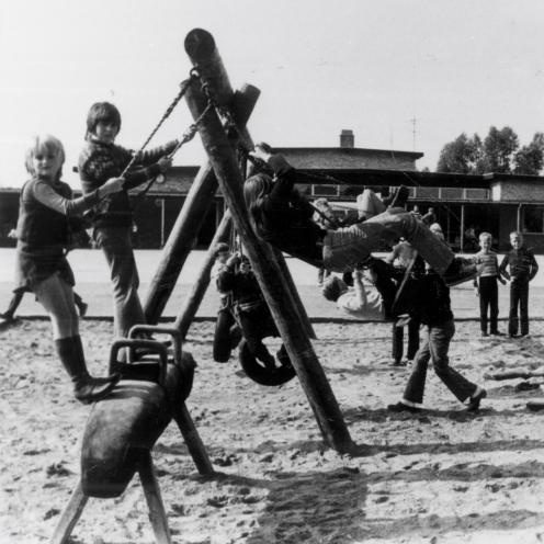 Sort-hvid foto af børn på gynger, som er opsat i en sandkasse i en skolegård.