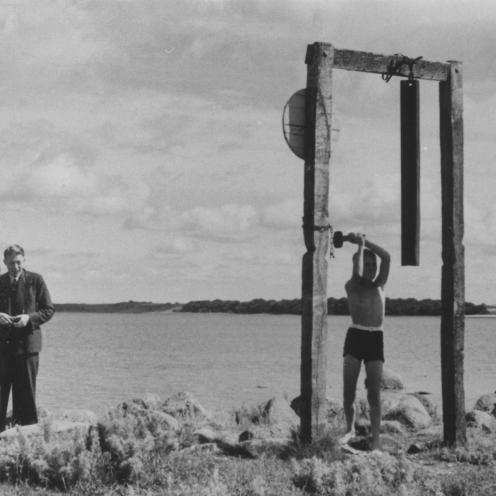 Sort-hvid foto af et barn der hamrer på en ophængt jernbaneskinne.