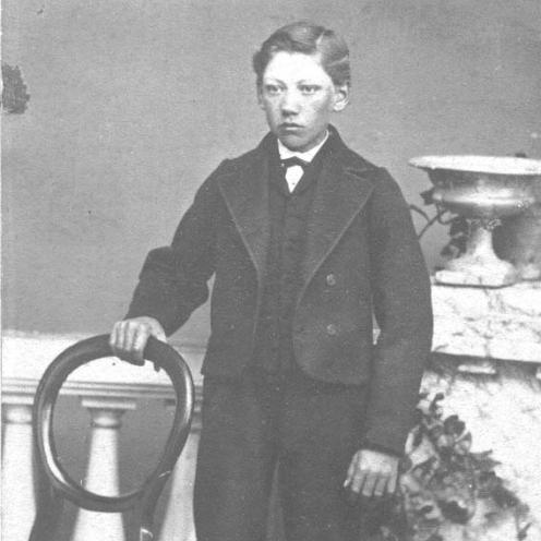 Sort-hvid portrætfoto af en dreng iklædt gammeldags tøj stående ved en stol.