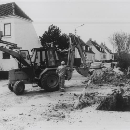 Sort-hvid foto af en gravemaskine og arbejdere ved et hul i asfalten.