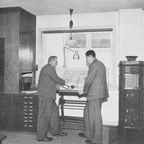 Sort-hvid foto af to mænd på en tegnestue ved en tegning af en maskine.