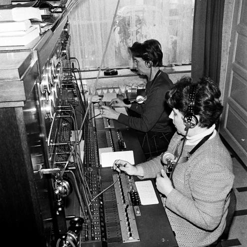 Kvindelige medarbejdere betjener et manuelt telefonbord med headset og stikforbindelser.