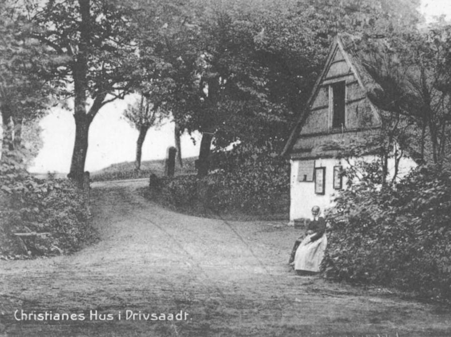 Sort-hvid foto: Facaden af Christianes Hus med Christiane, der sidder foran på en bænk.