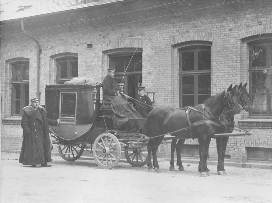 Sort-hvid foto: Postvognen / diligencen foran Jyderup Station. På bukken sidder Niels Jensen, i pels ved siden af står postkonduktør Jespersen og bag vognen postfuldmægtig Lynge. Datering: ca. 1905.