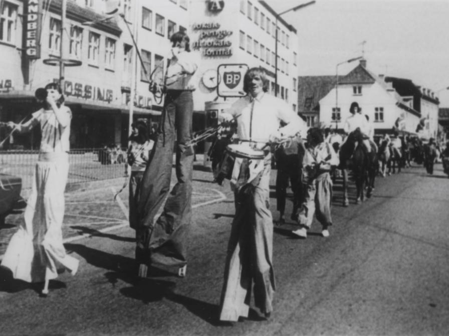 Sort-hvid foto: Parade af udklædte mennesker gående ad Ahlgade. Flere er på stylter.