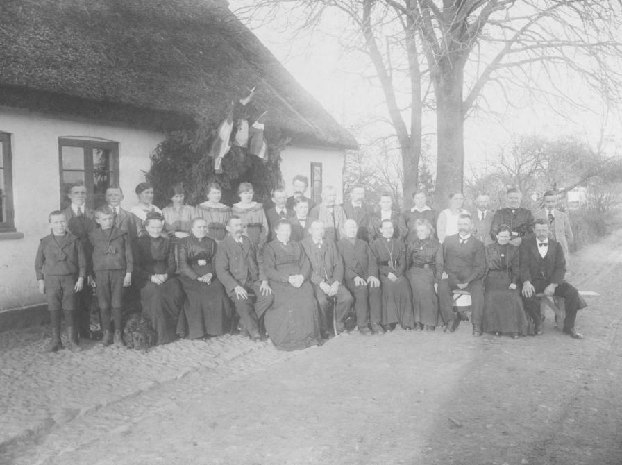 Sort-hvid foto: En flok mennesker opstillet til fotografering foran et stråtækt hus.