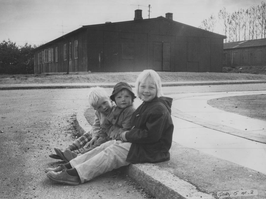 Sort-hvid foto af tre børn siddende på fortovet foran en sort træbygning.