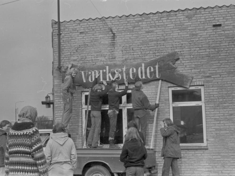 Sort-hvid foto af tre personer stående på ladet af en bil, der tager et skilt med teksten "Værkstedet" ned fra en murstensbygning.