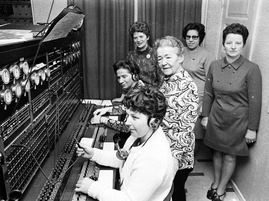 Kvindelige medarbejdere ved en manuel telefoncentral. Operatører arbejder ved centralbordet, mens kolleger står bagved.